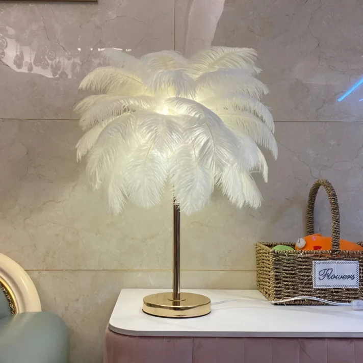 Ostrich feather table lamp with a gold metal base, white fluffy feather shade, placed on a white surface beside a woven basket, adding a luxurious, airy vibe.