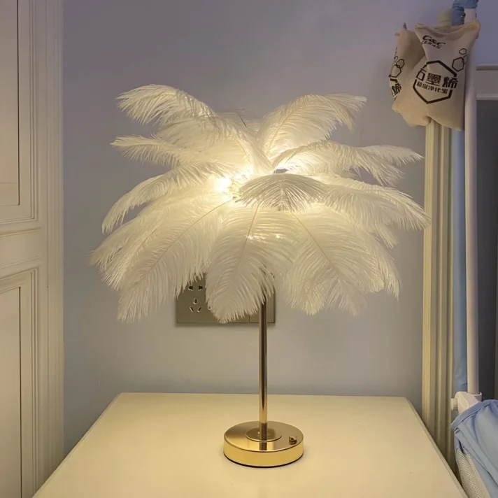 White ostrich feather table lamp with a sleek gold base, illuminated for a warm glow, from Verra Luxe Home's Rechargeable Ostrich Feather Table Lamp collection.