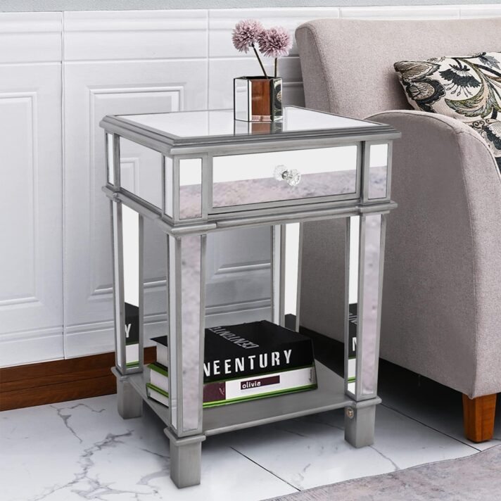Silver mirrored bedside table with a glass top and drawer, featuring a crystal-style handle, open shelf below, and chrome-finished frame, placed beside a beige sofa. Brand: Verra Luxe Home product: Silver Mirrored Bedside Table with Drawer & Open Shelf