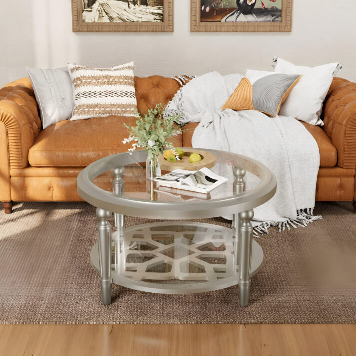 Verra Luxe Home round glass coffee table with storage featuring a champagne finish, two-tier design, and silver legs, placed in a living room with a tufted leather sofa.