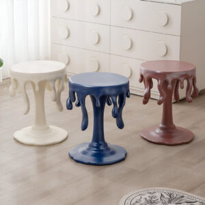 Three round Nordic-style resin stools in white, blue, and pink with dripping melted-edge details placed beside a white dresser in a minimal living space.