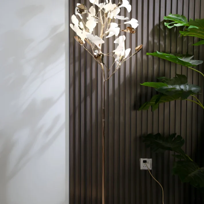 Nordic Ginkgo Leaf LED Floor Lamp by Verra Luxe Home in a warm gold finish with white ginkgo leaf accents, standing tall beside a dark paneled wall in a modern interior.