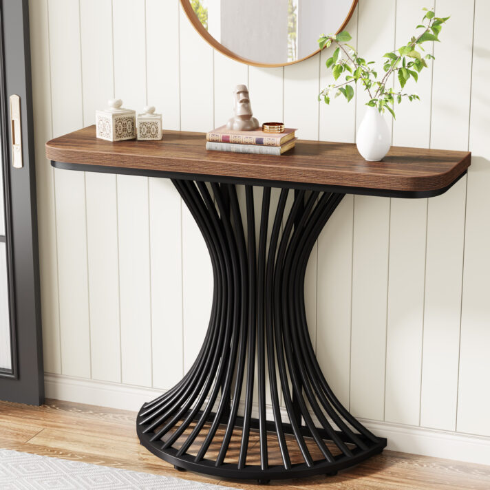 Verra Luxe Home industrial console table features a black curved metal base and a warm brown wood top, styled with decorative books, a ceramic vase, and greenery.