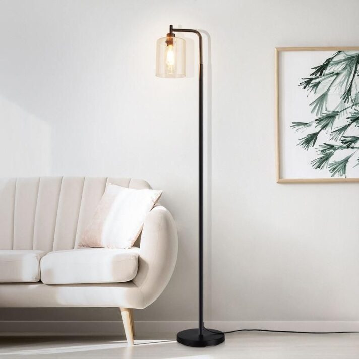 Elegant Nordic-inspired black metal LED floor lamp with a clear glass shade stands beside a light beige sofa in a bright, minimalist living room.
