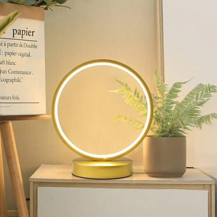Golden circular LED Sphere Table Lamp with glowing ring on a wooden dresser beside a potted fern, from Verra Luxe Home product imagery.
