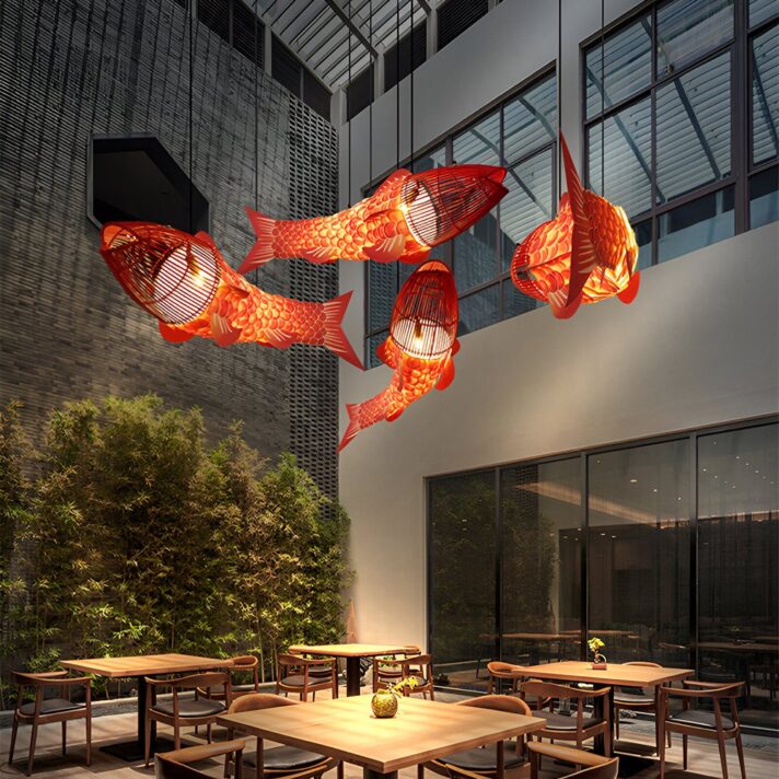 Red koi fish lanterns hang overhead in an outdoor courtyard with wooden tables and chairs, behind glass walls of a modern building.
