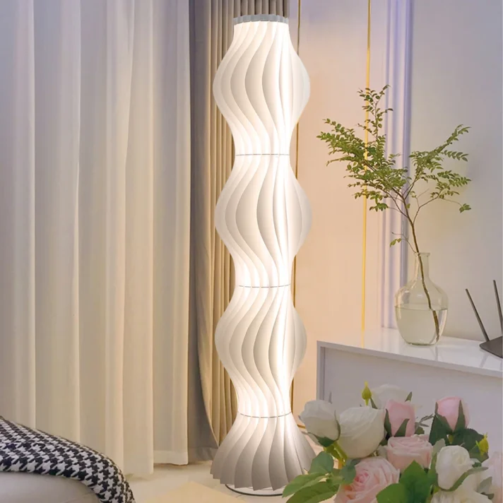 White sculptural wave-floor lamp from Verra Luxe Home, a tall modern lamp with curved panels beside beige curtains and a vase with greenery, enhancing luxe decor.