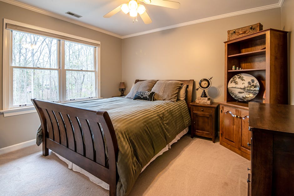 Beige bedroom with a dark wooden sleigh bed, matching nightstands, a tall cabinet, ceiling fan, and large window showing bare trees outside; decor includes a round plate and vintage items.