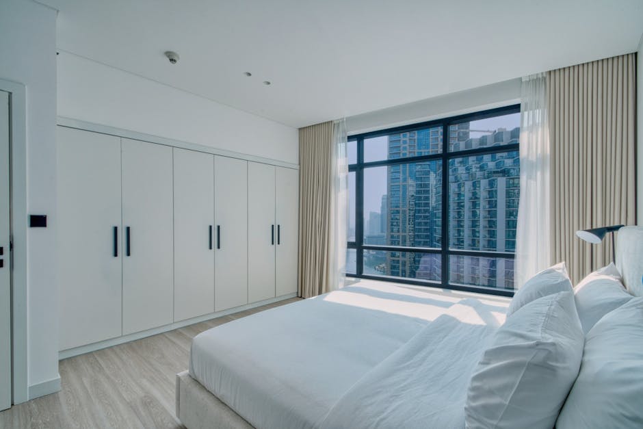 article-bbf2463b-9cba-41c2-b32d-e99d1b5d6ef2-featured White minimalist bedroom with built-in wardrobe, large window showing a city skyline, neutral tones, Verra Luxe Home interior styling.