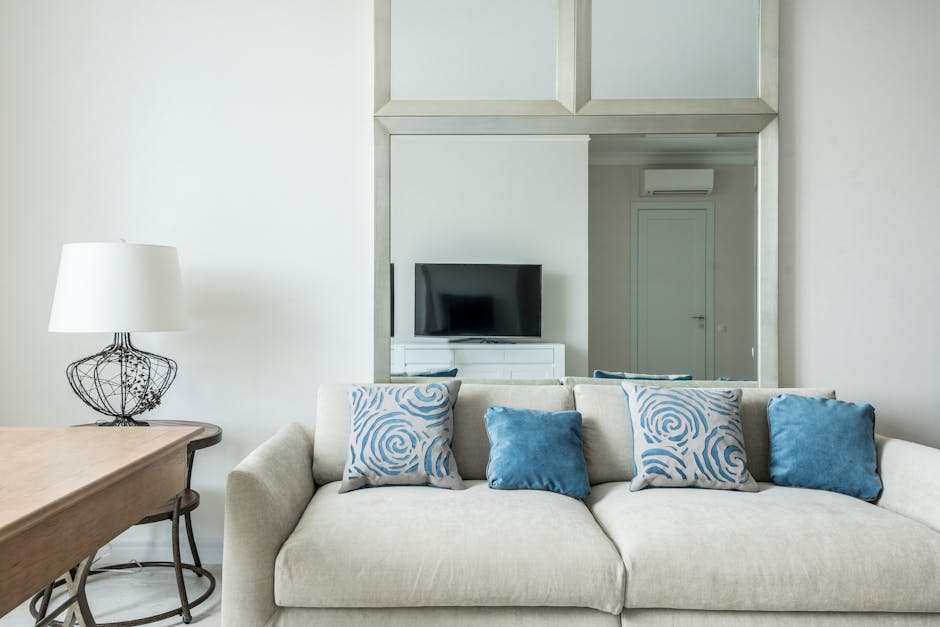 article-cb4fd4b4-27e6-461e-9fc9-f1a91ba42a73-featured Verra Luxe Home living room with a beige sofa adorned by blue patterned and solid cushions, a wooden table, and a large rectangular mirror reflecting a TV area.