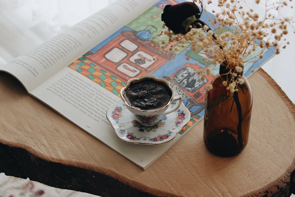 Brown glass vase with dried flowers beside an ornate porcelain coffee cup on a saucer atop an open magazine displaying colorful illustrated scenes
